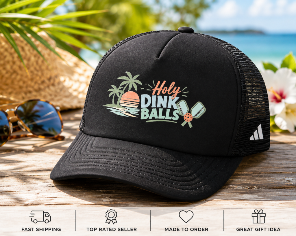 Black Holy Dink Balls pickleball trucker hat with retro beach sunset design displayed on a wooden table beside sunglasses at a tropical beach.